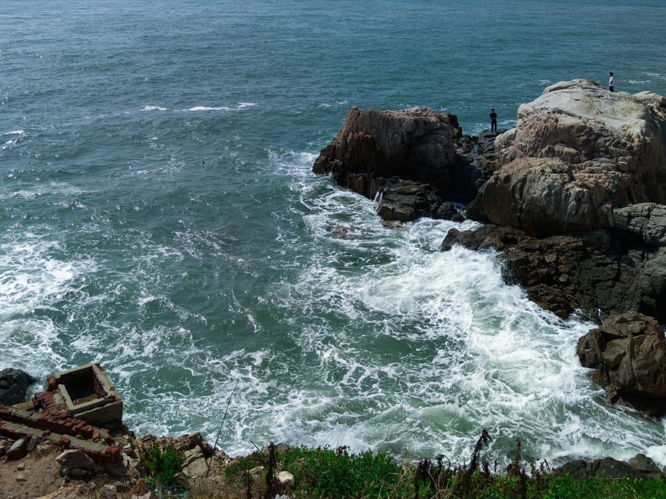 海浪拍打嶙峋礁石，岸边有人站立