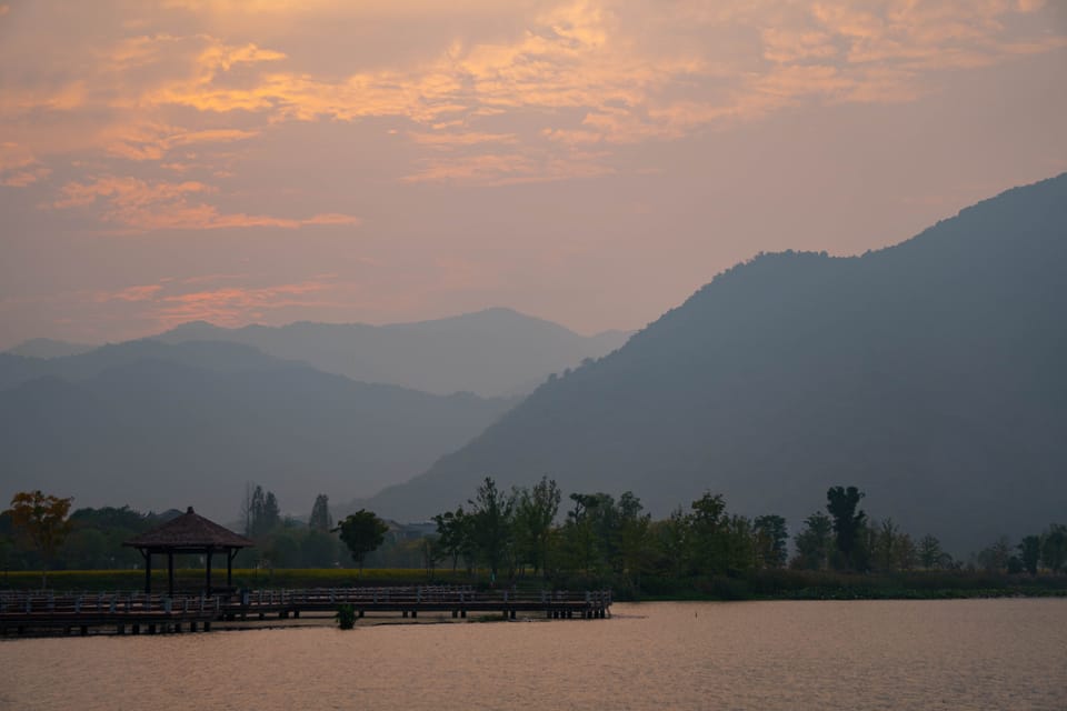暮色湖畔的木栈道与凉亭，背景是层叠青山与暖色天空