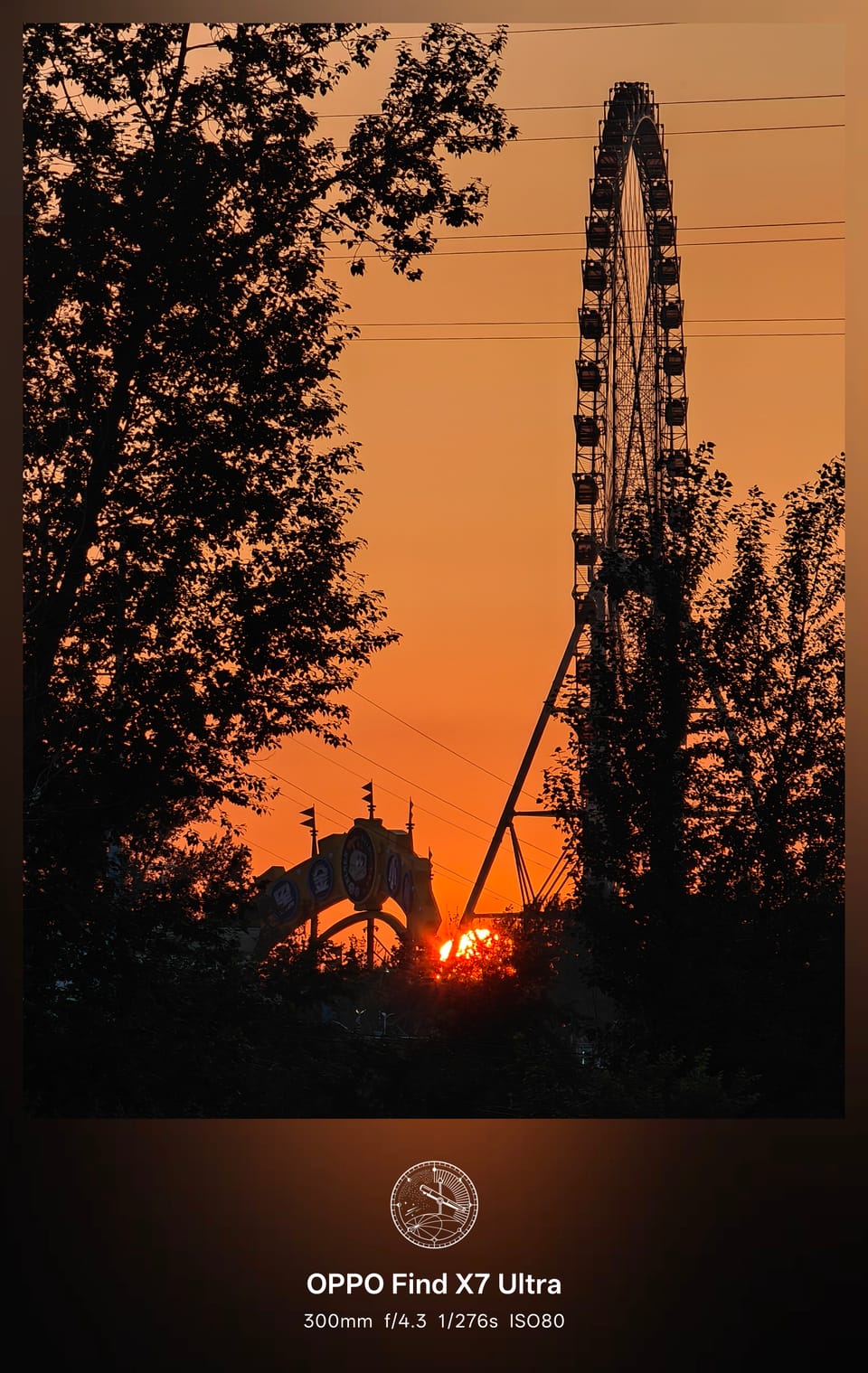 橙红天色中夕阳落在树影间，逆光的摩天轮与游乐场拱门轮廓