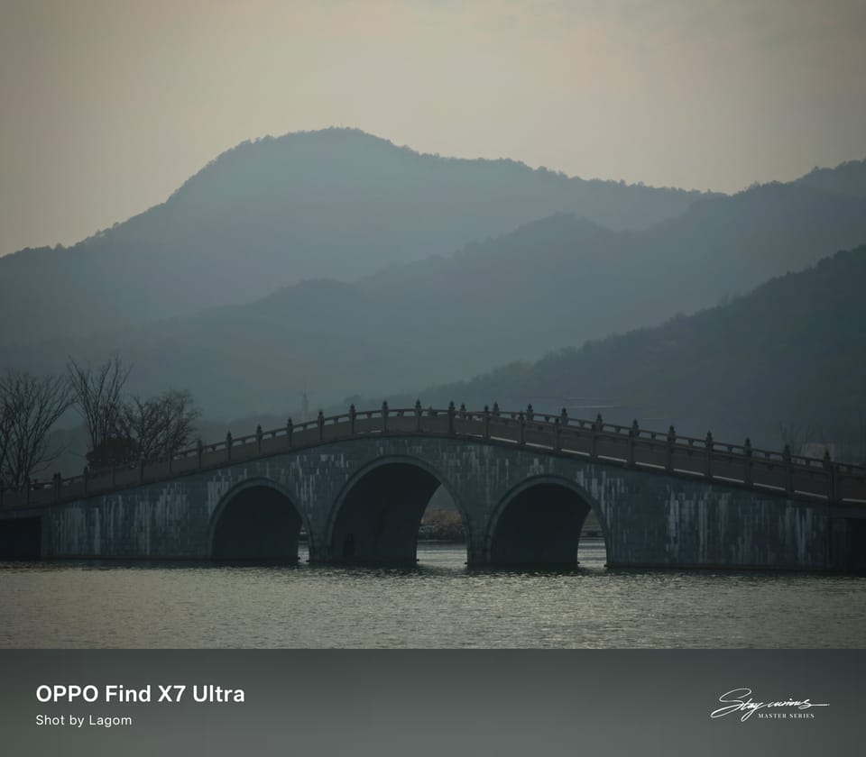 三孔石拱桥横跨平静水面，桥身倒映水中，背后是雾中层叠的山峦